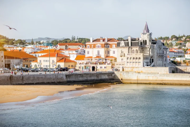 Scenic view of Cascais bay with historic palace, orange rooftops, beach, and people walking along the seafront promenade under clear sky.